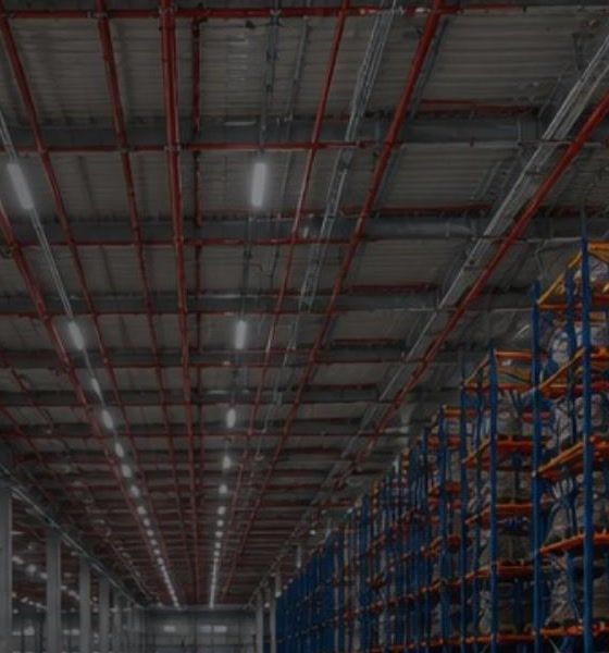 Allée d'entrepôt logistique : réseau sprinkler en acier peint courant sous la charpente, au-dessus de racks de stockage.